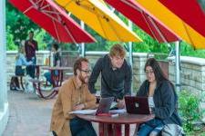 3 students look at a laptop on a picnic table under red 和 yellow umbrellas on the University of Denver campus. 